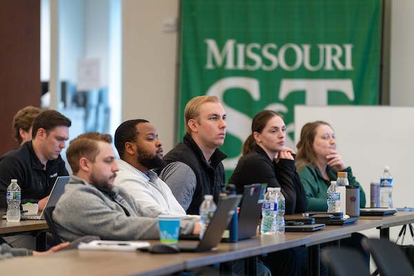 A group of focused individuals sit at a conference table with laptops and water bottles, listening to a presentation